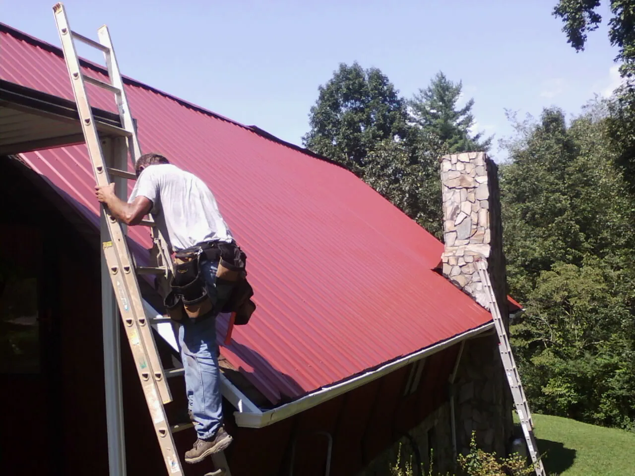 Roofer on a ladder beside a red metal roof and stone chimney — Soffit & Fascia Repair by Roofing Brothers in Canby